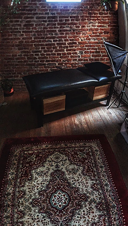 La camilla negra del estudio sobre piso de madera y una alfombra persa roja, junto a la pared de ladrillo.