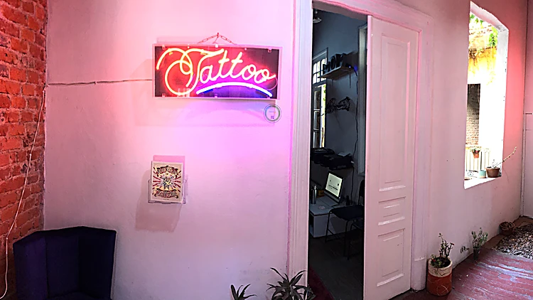 Cuarto del estudio con paredes rosas, el letrero de neón rojo que dice Tattoo y una puerta abierta al pasillo.