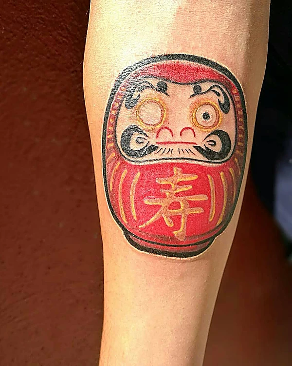 Daruma rojo a color con un kanji dorado, tatuado en la pantorrilla.