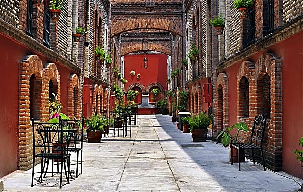 Pasillo de un edificio antiguo de ladrillo con arcos, macetas y bancas.