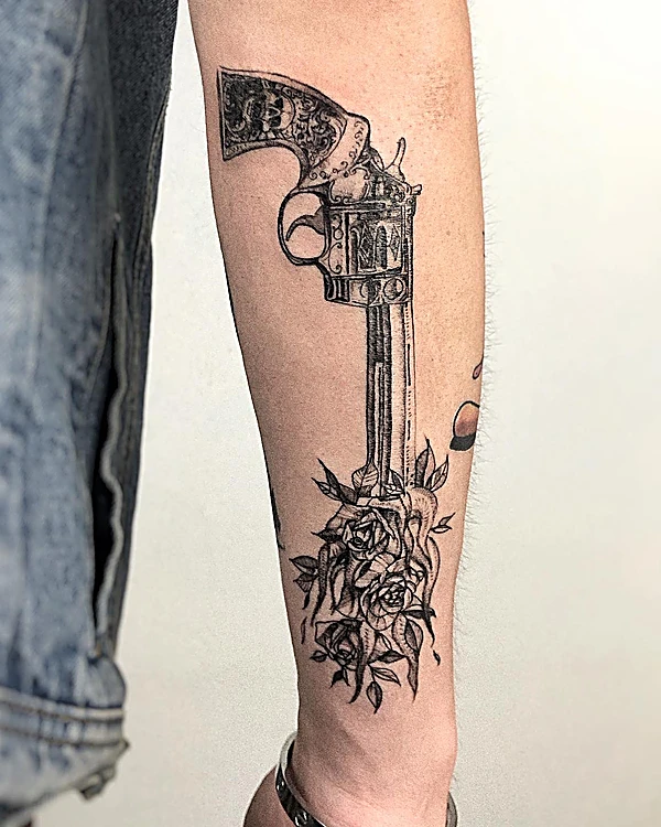 Revólver con rosas tatuado en negro y gris a lo largo del antebrazo.