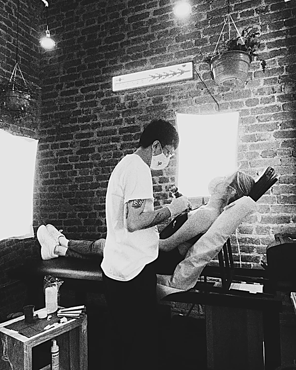 En blanco y negro: el tatuador con cubrebocas trabajando sobre la pierna de un cliente recostado, frente a la pared de ladrillo.