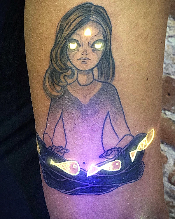 Tatuaje de una chica con tercer ojo y dos peces; los ojos y los peces brillan amarillos con luz UV.